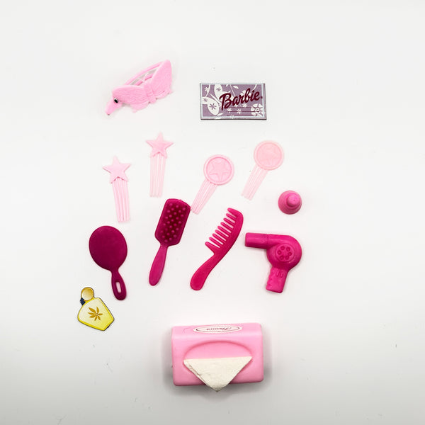 A set of Barbie-themed hair styling accessories in pink and magenta, including a hair clip, mirror, brushes, comb, hair dryer, hair tie, and a small yellow charm, arranged on a white background with a Barbie logo card.