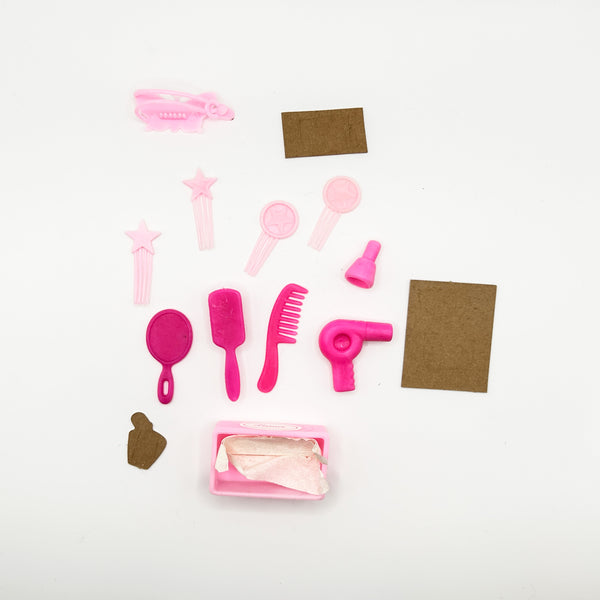 A set of miniature pink plastic doll hair accessories and styling tools, including a hair clip, star-shaped hairpins, a comb, a hairbrush, a hair dryer, and a small mirror, arranged on a white background with two brown paper labels.