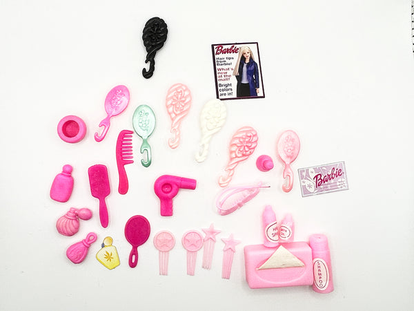 A mixed lot of vintage Barbie doll hair styling and vanity accessories arranged on a white background, featuring various pink brushes, combs, a miniature toy hairdryer, accessory bottles, a small paper Barbie styling booklet, and a black hook brush