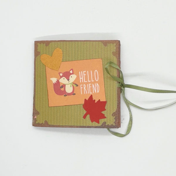 Handmade mini autumn scrapbook album with an orange and green striped patterned cover, featuring a cartoon fox illustration, a die-cut red maple leaf, a gold heart, and the text "HELLO FRIEND." A green satin ribbon closure extends from the side of the book, which sits on a wooden shelf against a brick background.
