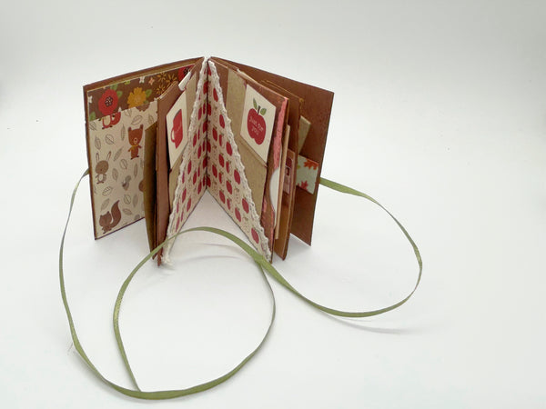 Handmade mini autumn scrapbook album with an orange and green striped patterned cover, featuring a cartoon fox illustration, a die-cut red maple leaf, a gold heart, and the text "HELLO FRIEND." A green satin ribbon closure extends from the side of the book, which sits on a wooden shelf against a brick background.
