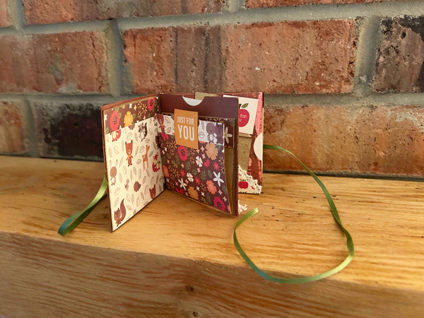Handmade mini autumn scrapbook album with an orange and green striped patterned cover, featuring a cartoon fox illustration, a die-cut red maple leaf, a gold heart, and the text "HELLO FRIEND." A green satin ribbon closure extends from the side of the book, which sits on a wooden shelf against a brick background.
