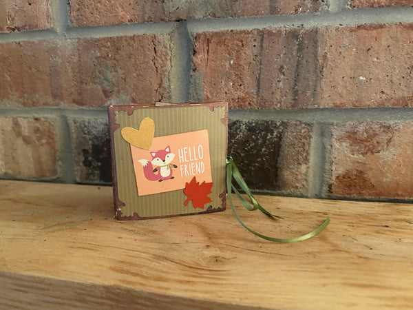 Handmade mini autumn scrapbook album with an orange and green striped patterned cover, featuring a cartoon fox illustration, a die-cut red maple leaf, a gold heart, and the text "HELLO FRIEND." A green satin ribbon closure extends from the side of the book, which sits on a wooden shelf against a brick background.

