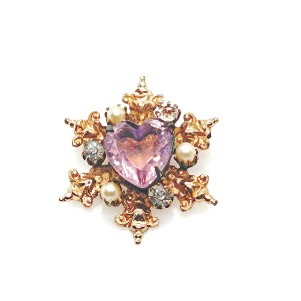 A gold-toned brooch with a central heart-shaped, light purple gemstone. The brooch is surrounded by ornate gold filigree, small pearls, and clear crystals, creating a vintage, elegant design.
