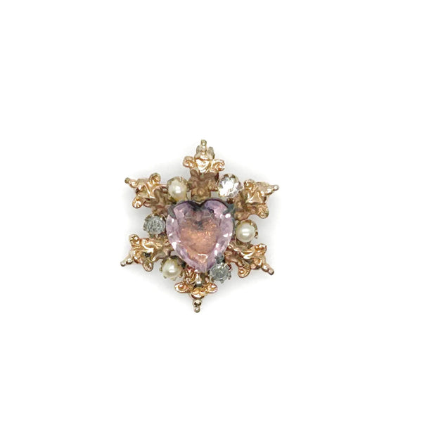A gold-toned brooch with a central heart-shaped, light purple gemstone. The brooch is surrounded by ornate gold filigree, small pearls, and clear crystals, creating a vintage, elegant design.