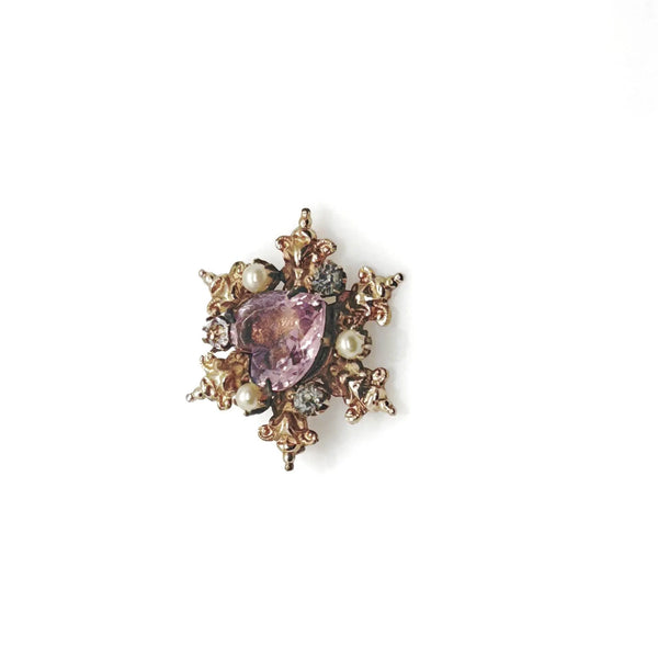 A gold-toned brooch with a central heart-shaped, light purple gemstone. The brooch is surrounded by ornate gold filigree, small pearls, and clear crystals, creating a vintage, elegant design.