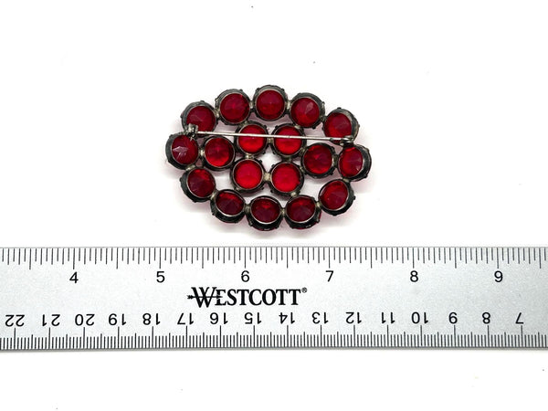 A vintage-style oval brooch with red faceted rhinestones in silver-toned settings, shown above a Westcott ruler for scale on a white background.