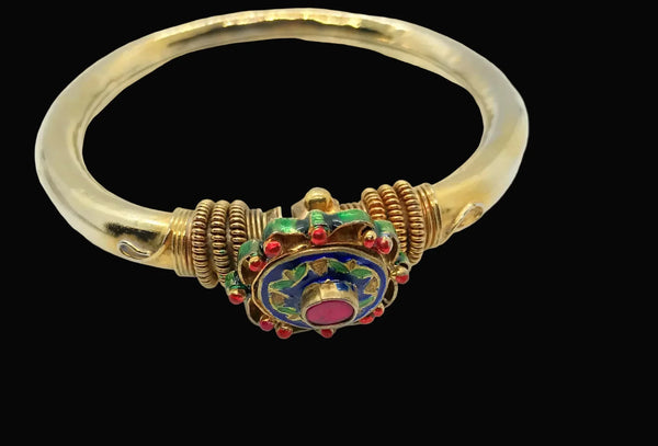 A gold bangle bracelet featuring a central decorative clasp with blue enamel, red stones, and green accents, shown against a dark gray background.