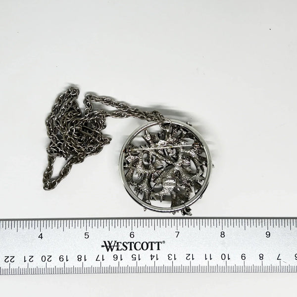 This striking, rounded, and very detailed brooch pendant makes a beautiful necklace. It measures 2" across and comes on a 24" chain with a secure fold-over clasp. The pendant features a strong, eye-catching frame that showcases a delicate floral motif, complete with blooms and leaves