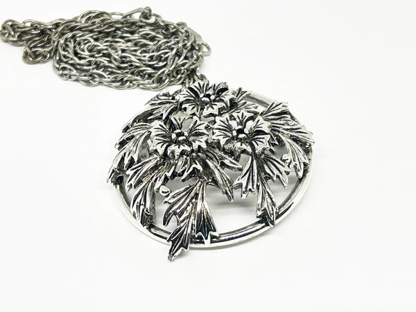 This striking, rounded, and very detailed brooch pendant makes a beautiful necklace. It measures 2" across and comes on a 24" chain with a secure fold-over clasp. The pendant features a strong, eye-catching frame that showcases a delicate floral motif, complete with blooms and leaves