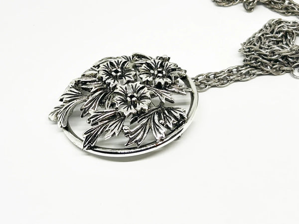 This striking, rounded, and very detailed brooch pendant makes a beautiful necklace. It measures 2" across and comes on a 24" chain with a secure fold-over clasp. The pendant features a strong, eye-catching frame that showcases a delicate floral motif, complete with blooms and leaves