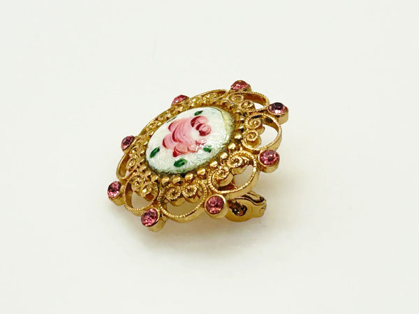 Take a look at this vintage costume jewelry brooch featuring a hand-painted pink rose on a white enamel or ceramic base, set within an ornate gold-tone filigree frame accented with pink rhinestones. The center showcases a pink rose, with green dots accenting the perimeter. The setting and metalwork around the outside are particularly lovely. It measures 1" across.