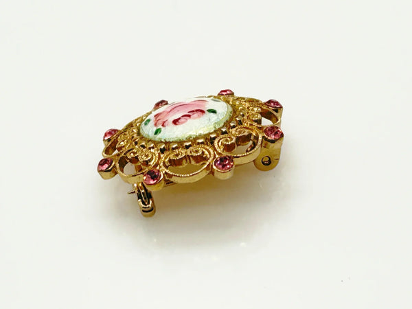 Take a look at this vintage costume jewelry brooch featuring a hand-painted pink rose on a white enamel or ceramic base, set within an ornate gold-tone filigree frame accented with pink rhinestones. The center showcases a pink rose, with green dots accenting the perimeter. The setting and metalwork around the outside are particularly lovely. It measures 1" across.