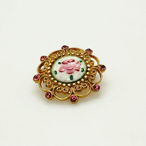 Take a look at this vintage costume jewelry brooch featuring a hand-painted pink rose on a white enamel or ceramic base, set within an ornate gold-tone filigree frame accented with pink rhinestones. The center showcases a pink rose, with green dots accenting the perimeter. The setting and metalwork around the outside are particularly lovely. It measures 1" across.