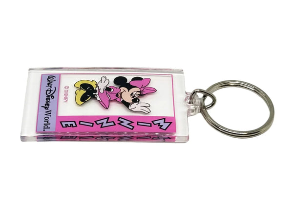 This Minnie Mouse keychain is too cute. It is made of clear acrylic, and shows Minnie Mouse in a pink dress, a pink bow, white gloves and yellow shoes. She has a big smile and is waving. The letters on the left side read "Minnie" in lavender with black border, on a bright pink background. Below Minnie is a purple banner with "Walt Disney World" in black