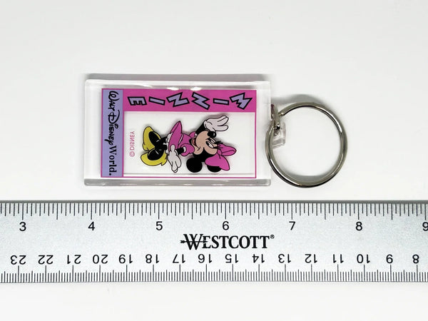 This Minnie Mouse keychain is too cute. It is made of clear acrylic, and shows Minnie Mouse in a pink dress, a pink bow, white gloves and yellow shoes. She has a big smile and is waving. The letters on the left side read "Minnie" in lavender with black border, on a bright pink background. Below Minnie is a purple banner with "Walt Disney World" in black