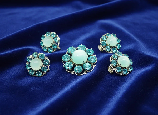 This is a hard-to-find set featuring a brooch, a pair of scatter pins, and screw-back earrings. The design showcases round, frosted light turquoise cabochons. Each features a center moonglow stone surrounded by bright, faceted, slightly deeper blue stones, all prong-set. 