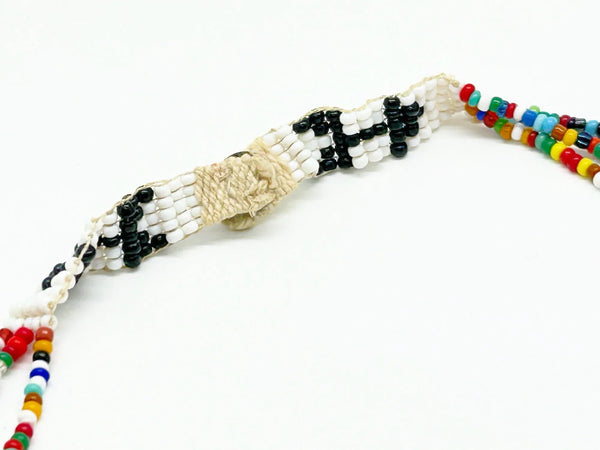 This striking Native American beaded necklace features a beautifully detailed figure adorned with two feathers in flowing black hair, dressed in a brown tunic and red skirt with black shoes. The design showcases a white upper background that transitions to a vibrant turquoise-teal green below, creating visual depth and interest. 