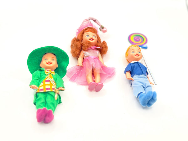 Three loose vintage 1999 Mattel Barbie Kelly Club mini dolls from The Wizard of Oz line arranged on a white background: Tommy as Mayor Munchkin in a green felt top hat and jacket on the left, Kelly as Lullaby Munchkin in a pink conical hat and tutu in the center, and Tommy as Lollipop Munchkin holding a colorful swirl lollipop on the right