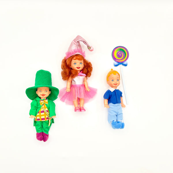 Three loose vintage 1999 Mattel Barbie Kelly Club mini dolls from The Wizard of Oz line arranged on a white background: Tommy as Mayor Munchkin in a green felt top hat and jacket on the left, Kelly as Lullaby Munchkin in a pink conical hat and tutu in the center, and Tommy as Lollipop Munchkin holding a colorful swirl lollipop on the right