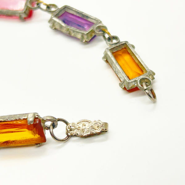 Close-up of a vintage-style gemstone bracelet featuring rectangular amber-orange, purple, and pink crystals set in silver-tone metal, with an ornate clasp detail on a white background.