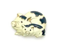 A vintage DKI gold-tone enamel brooch shaped like two snuggling black and white spotted pigs on a white background.