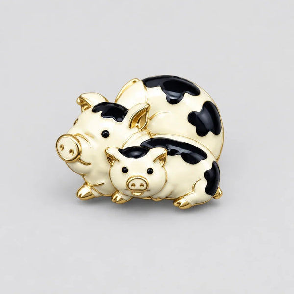 A whimsical vintage gold-tone brooch featuring a stacked pair of cream enamel pigs decorated with smooth black cow-print spots. The playful design highlights polished gold-plated snouts, ears, and hooves, cleanly photographed against a neutral light gray background.