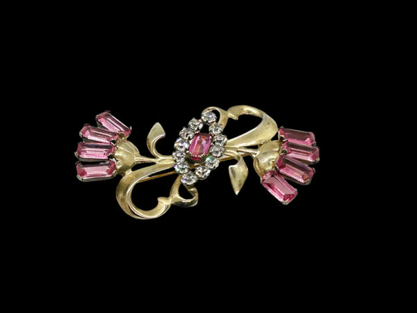 This stunning vintage brooch exudes the essence of classic style. Featuring delicate flowers adorned pink, faceted open back stones as petals, and with leaf accents on either end, the centerpiece boasts a striking marquise-shaped design embellished with dazzling white round stones and a magnificent pink emerald cut stone, mirroring those in the flowers.