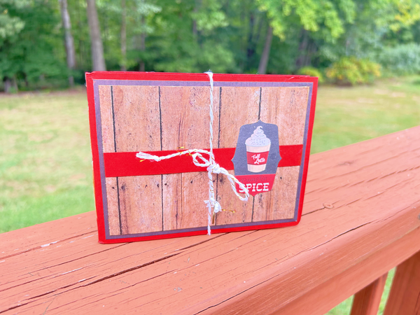 Handmade autumn mini scrapbook album with a rustic brown wood-plank pattern cover, wrapped with a bright orange paper band. The cover features a dark shield-shaped tag with an illustration of a "Fall Latte" whipped cream drink and red text that reads "Pumpkin Spice."