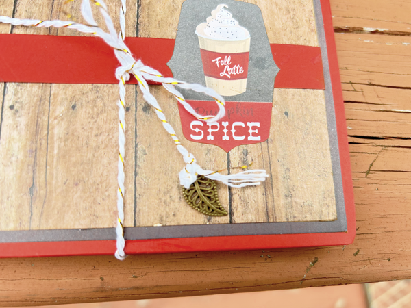 A close-up of a decorative on a fall themed mini album package wrapped with white and yellow twine, featuring a rustic wood-grain design, a red stripe, a coffee cup graphic that says 'Fall Little,' and the word 'SPICE' on a wooden surface.