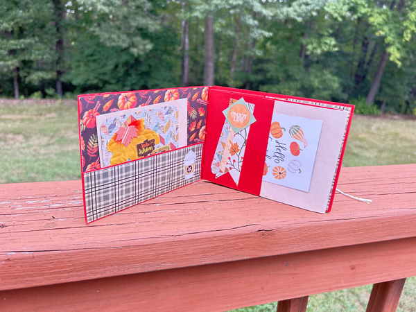 Handmade autumn mini scrapbook album with a rustic brown wood-plank pattern cover, wrapped with a bright orange paper band. The cover features a dark shield-shaped tag with an illustration of a "Fall Latte" whipped cream drink and red text that reads "Pumpkin Spice."