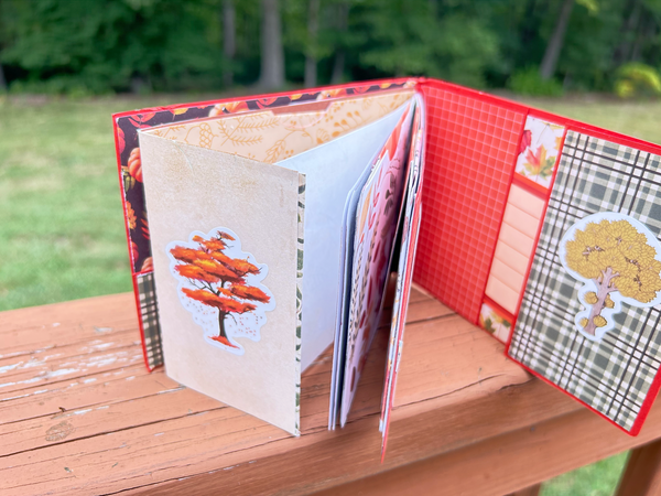 Handmade autumn mini scrapbook album with a rustic brown wood-plank pattern cover, wrapped with a bright orange paper band. The cover features a dark shield-shaped tag with an illustration of a "Fall Latte" whipped cream drink and red text that reads "Pumpkin Spice."