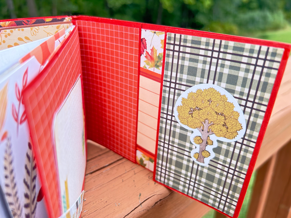 Handmade autumn mini scrapbook album with a rustic brown wood-plank pattern cover, wrapped with a bright orange paper band. The cover features a dark shield-shaped tag with an illustration of a "Fall Latte" whipped cream drink and red text that reads "Pumpkin Spice."