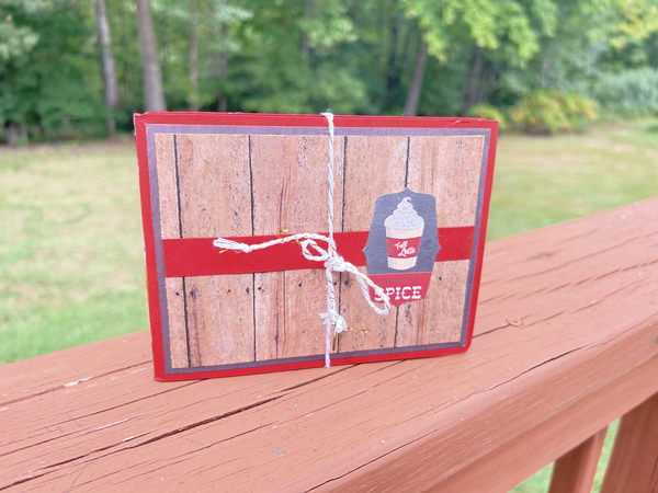 Handmade autumn mini scrapbook album with a rustic brown wood-plank pattern cover, wrapped with a bright orange paper band. The cover features a dark shield-shaped tag with an illustration of a "Fall Latte" whipped cream drink and red text that reads "Pumpkin Spice."
