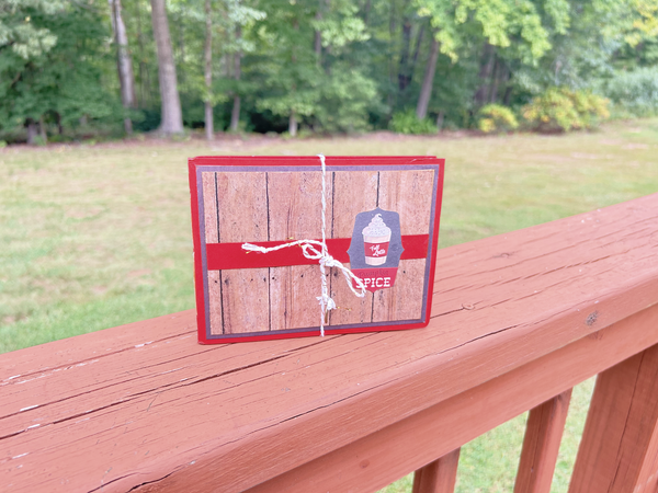 Handmade autumn mini scrapbook album with a rustic brown wood-plank pattern cover, wrapped with a bright orange paper band. The cover features a dark shield-shaped tag with an illustration of a "Fall Latte" whipped cream drink and red text that reads "Pumpkin Spice."