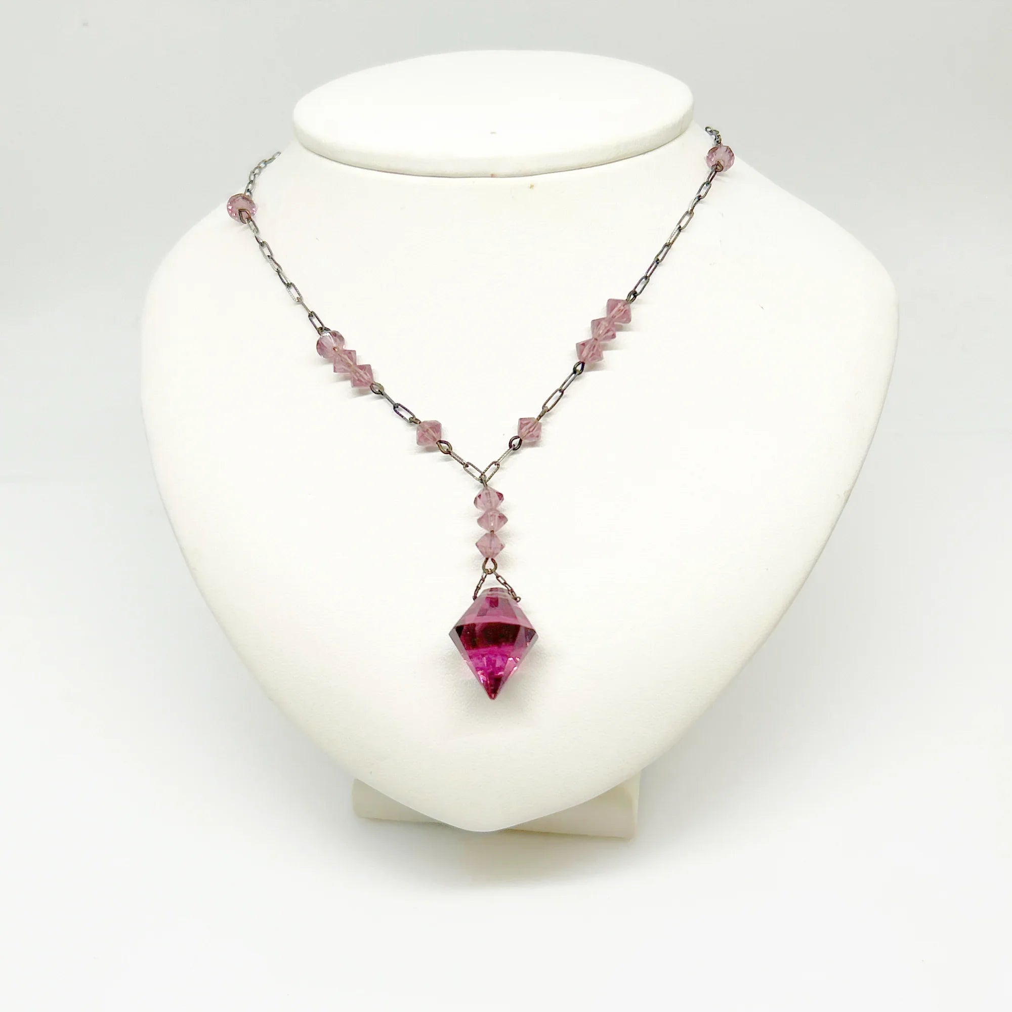 This stunning vintage crystal necklace showcases clusters of deep purple bicone beads, interspersed with sleek paper clip chains. It culminates in a magnificent faceted diamond-shaped stone. Measuring 16.5 inches in length, with a drop of 1.5 inches from the center, this piece exudes elegance and sophistication.