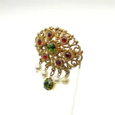 Product photo of a vintage antique-gold filigree pin. The central oval frame holds bright green and red glass gems. Six small dangling charms hang from the bottom, including five creamy white pearls and one round green rhinestone.