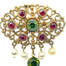 Product photo of a vintage antique-gold filigree pin. The central oval frame holds bright green and red glass gems. Six small dangling charms hang from the bottom, including five creamy white pearls and one round green rhinestone.