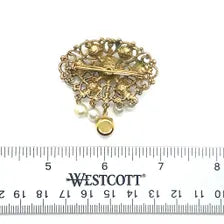 Product photo of a vintage antique-gold filigree pin. The central oval frame holds bright green and red glass gems. Six small dangling charms hang from the bottom, including five creamy white pearls and one round green rhinestone.