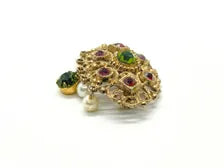 Product photo of a vintage antique-gold filigree pin. The central oval frame holds bright green and red glass gems. Six small dangling charms hang from the bottom, including five creamy white pearls and one round green rhinestone.