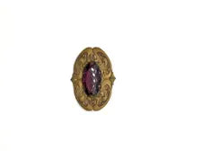 Antique Art Nouveau sash pin brooch crafted from embossed gold-tone brass with swirling whiplash foliage curves, featuring a large prong-set oval faceted purple amethyst glass gemstone center.