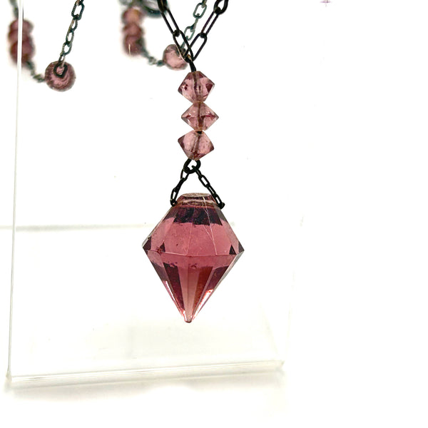 A vintage necklace featuring clusters of deep purple bicone crystal beads on a dark-toned paperclip chain, culminating in a large, heavily faceted diamond-shaped purple crystal drop pendant