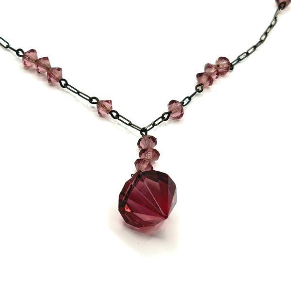 A vintage necklace featuring clusters of deep purple bicone crystal beads on a dark-toned paperclip chain, culminating in a large, heavily faceted diamond-shaped purple crystal drop pendant