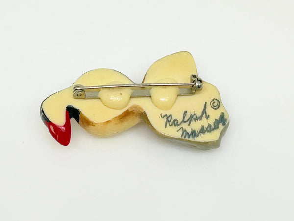 This is a brooch by artist Ralph Massey, who is acclaimed for many types of artwork- acrylic, performance, resin and more. This brooch is resin, hand made and hand painted in white, black, red-orange and gray. It depicts a penguin standing on rock, and measures about 1.75” inches tall.