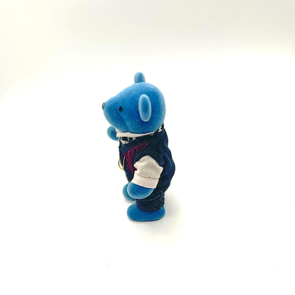 A small blue teddy bear figurine wearing a white shirt, dark vest, striped tie, and dark trousers, standing against a plain white background.