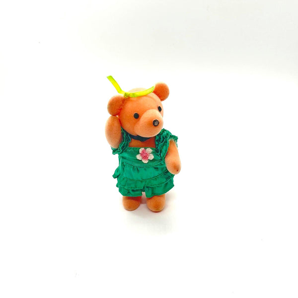 A small orange teddy bear figurine wearing a green ruffled dress with a pink flower accent and a yellow leaf on its head, standing against a plain white background.
