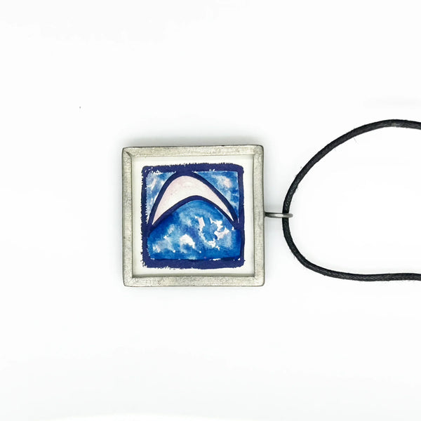 This is a modern inspiration in the form of a square pendant centering around the quote "Every possibility begins with the courage to imagine" by Mary Anne Radmacher. The quote is written in black ink, on a white background, and framed in silver metal. The reverse features a blue night sky background with a quarter moon and star. 