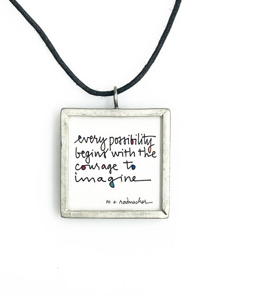 This is a modern inspiration in the form of a square pendant centering around the quote "Every possibility begins with the courage to imagine" by Mary Anne Radmacher. The quote is written in black ink, on a white background, and framed in silver metal. The reverse features a blue night sky background with a quarter moon and star. 