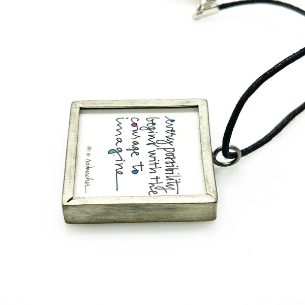 This is a modern inspiration in the form of a square pendant centering around the quote "Every possibility begins with the courage to imagine" by Mary Anne Radmacher. The quote is written in black ink, on a white background, and framed in silver metal. The reverse features a blue night sky background with a quarter moon and star. 