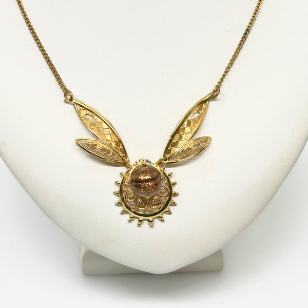 A gold pendant necklace displayed on a white jewelry bust. The necklace features a central oval amber-brown stone surrounded by a sunburst pattern, with two textured leaf-like gold wings extending upward from the pendant on a fine gold chain.